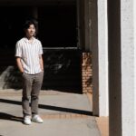 A young man in a button up shirt standing in the sun inside a hallway of a college campus.