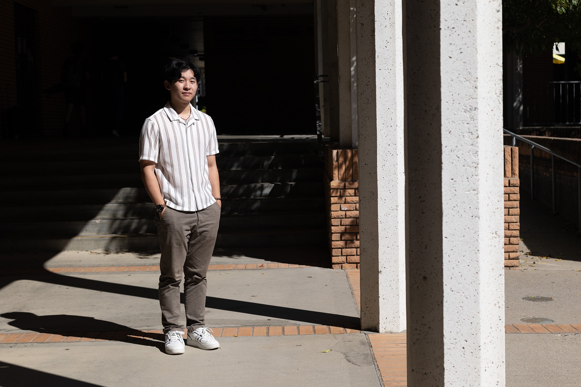 A young man in a button up shirt standing in the sun inside a hallway of a college campus.