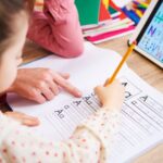 Teacher helping young child write the letter A on a worksheet in a notebook.