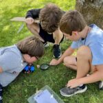 Vermont afterschool: Three young children in summer clithes crouch close together on grass in a circle playing a game.