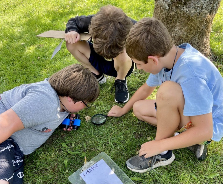 Vermont afterschool: Three young children in summer clithes crouch close together on grass in a circle playing a game.